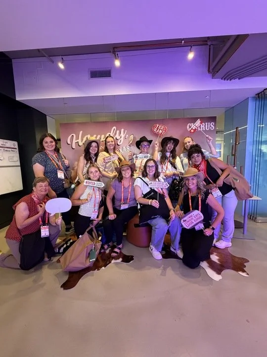 Group of women in country-themed costumes at a Texas themed event with a backdrop that reads 'Howdy Texas.' They are holding signs and props, and some are wearing cowboy hats, with a cowhide rug on the floor.