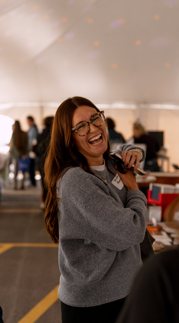 Woman with glasses singing and holding a microphone at an event under a tent.