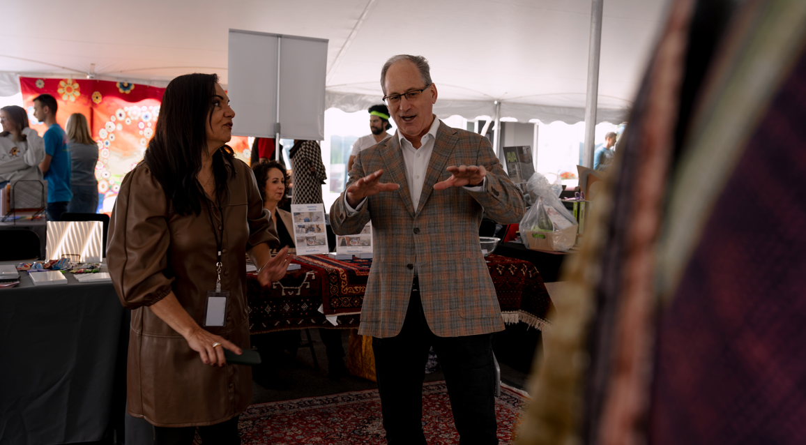 Two people engaged in conversation at an indoor event, with booths and other attendees visible in the background.