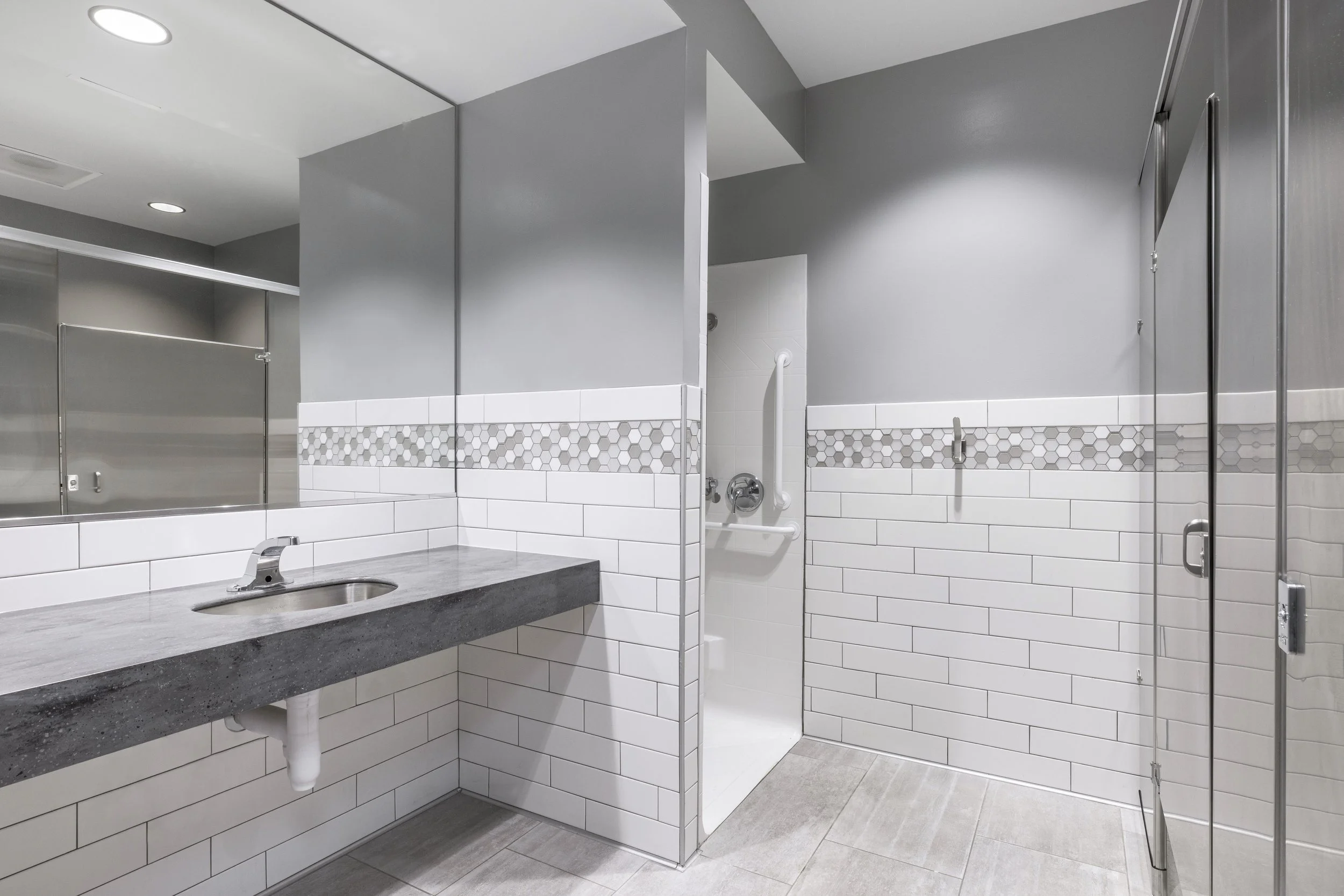 Modern indoor bathroom area with gray painted walls, white tiled lower walls with hexagonal accents, a gray countertop with a sink, a large mirror, and a shower with clear glass door on the right.