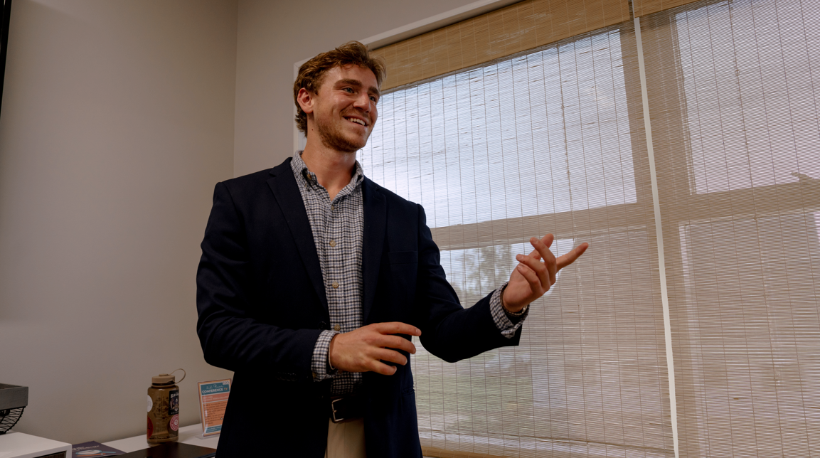 A young man in a blazer and checkered shirt smiling and gesturing with his hand in a bright room with window blinds.