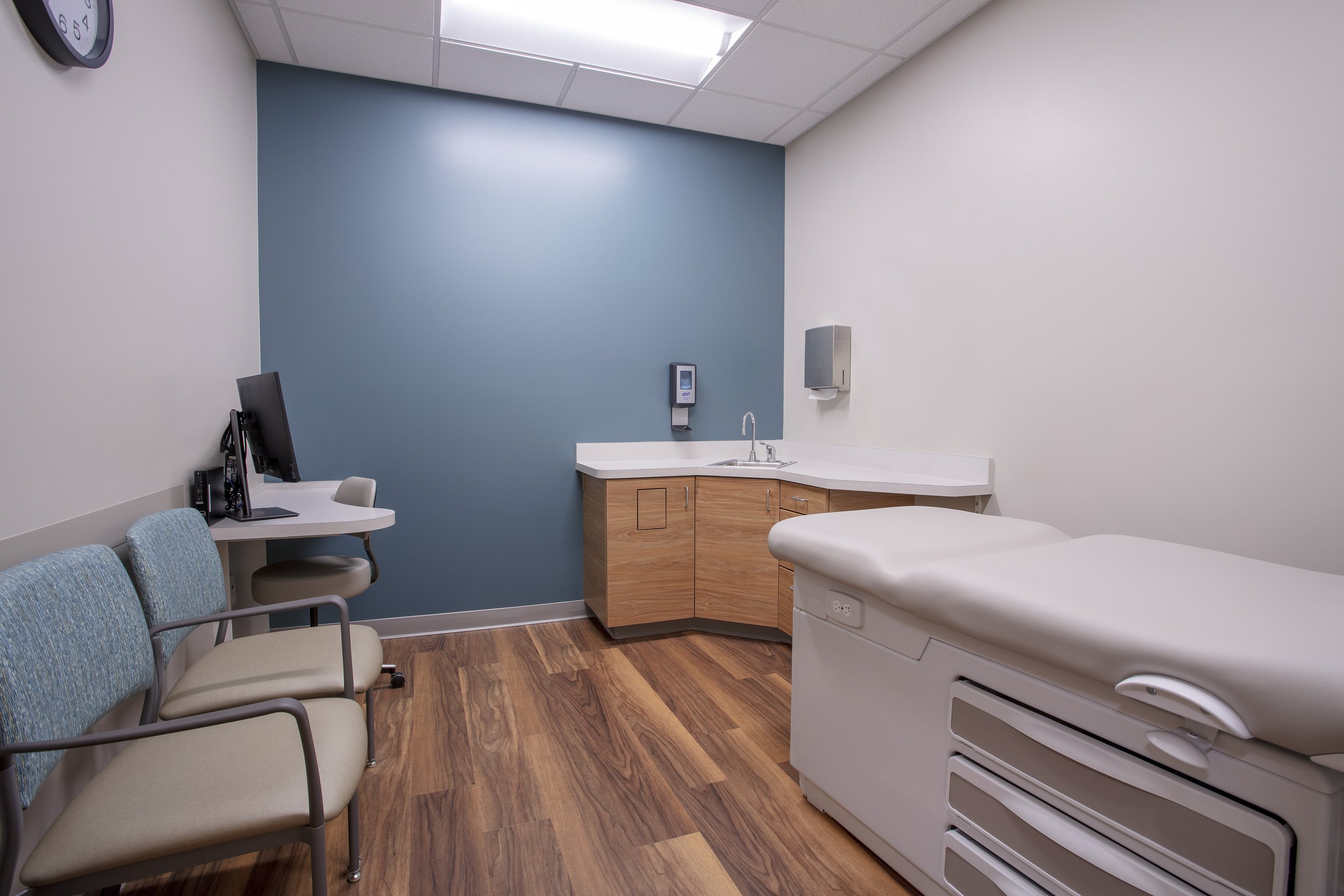 A hospital room with a light blue accent wall, a bed, a patient monitor, a computer on a small desk, a row of chairs, and medical supplies including soap and paper towel dispensers.