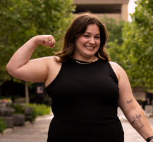 A smiling woman with shoulder-length brown hair in a black sleeveless top, flexing her right arm outdoors with trees and buildings in the background.