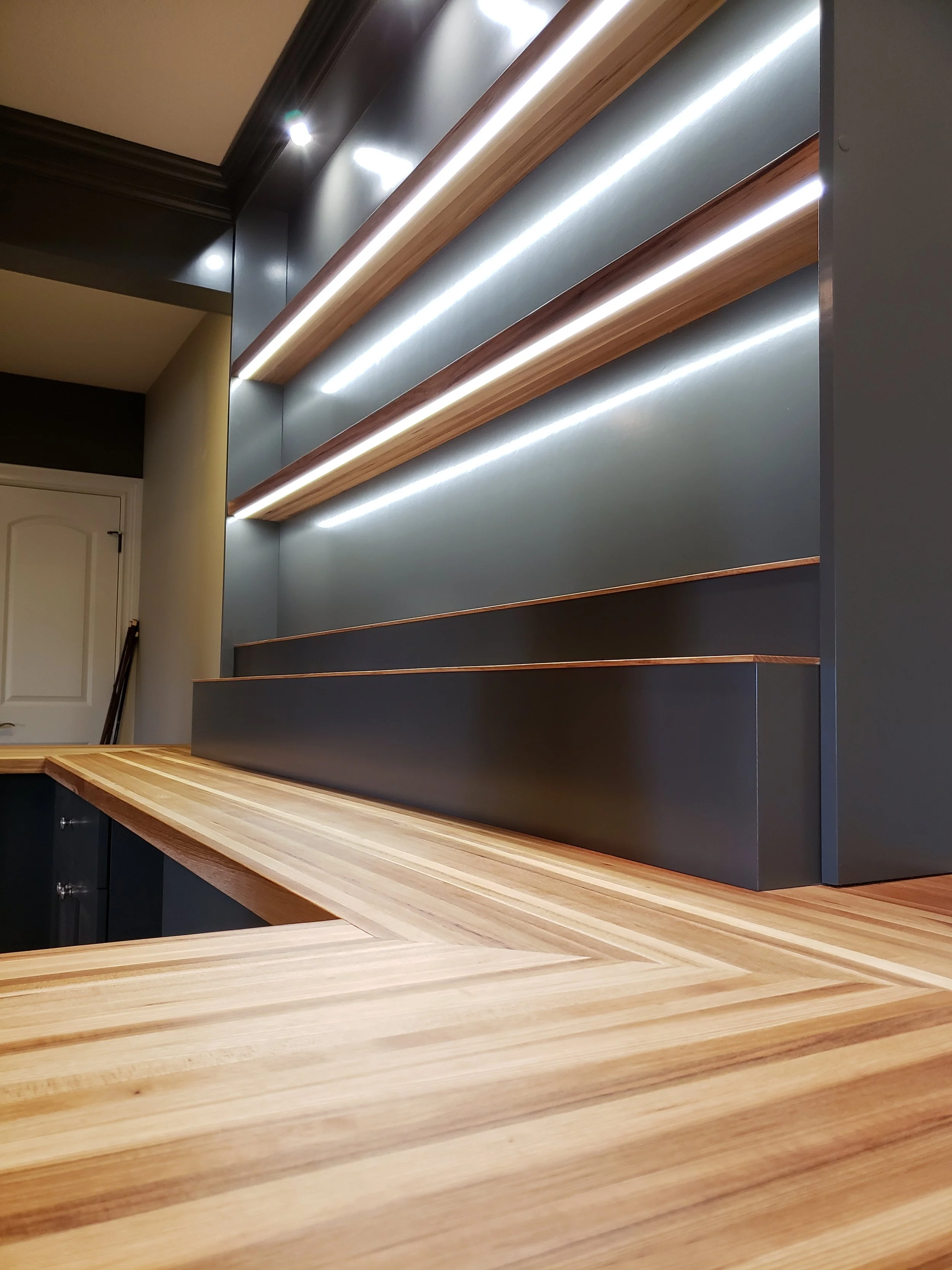 Wooden countertop with a dark gray/blue backsplash with wooden shelves and under-shelf lighting in a modern kitchen or bar area.