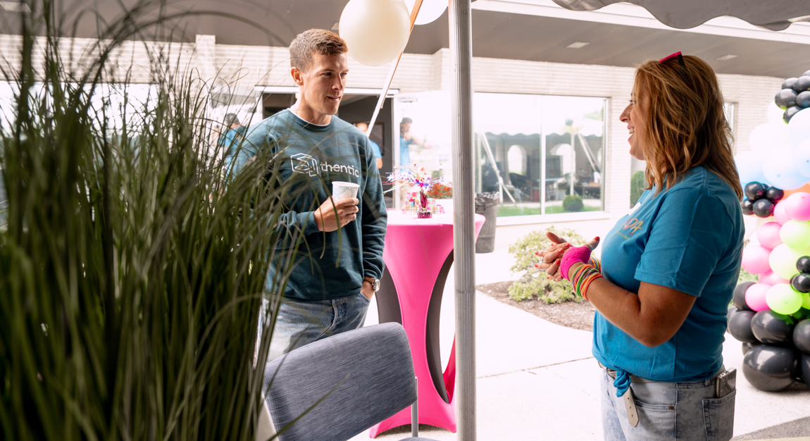 A young man and a woman are talking at an outdoor event. The man is holding a cup and wearing a dark blue shirt with 'Authentic' written on it. The woman, wearing a similar blue shirt, has colorful bracelets and pink gloves. They are standing near a 