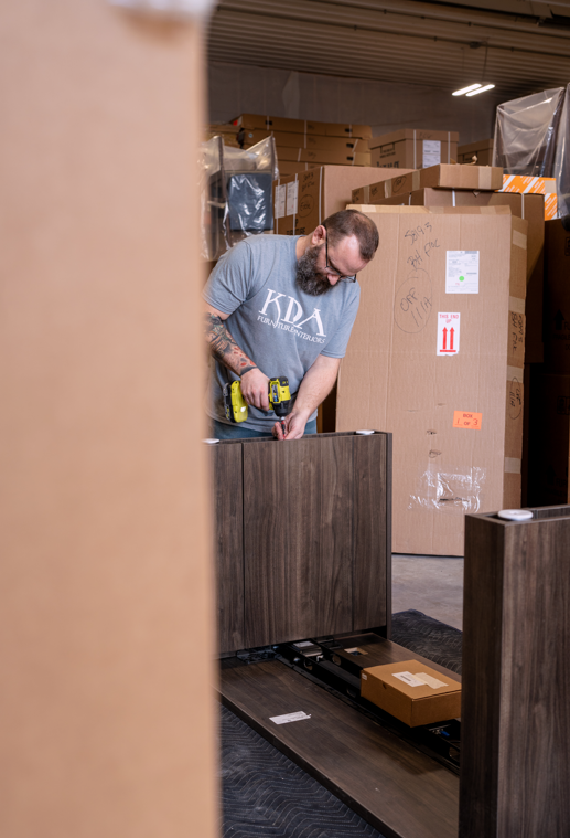 A man wearing a gray T-shirt with DEA Furniture logo is using a cordless drill to assemble a piece of furniture in a warehouse filled with stacked cardboard moving boxes.