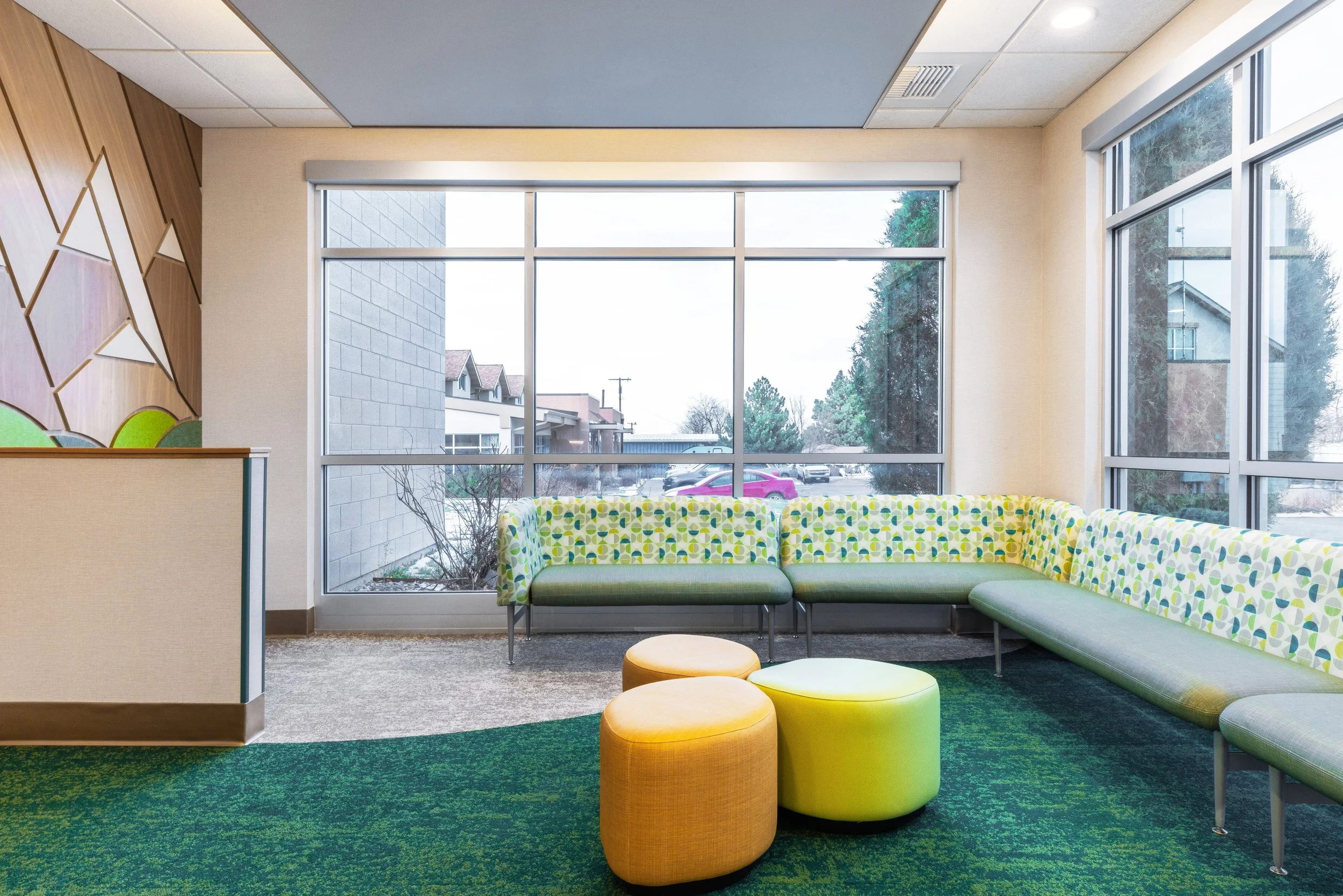 Interior view of a waiting or lounge area with large windows, a patterned green and yellow sectional sofa, three colorful ottomans, and a decorative wooden wall with geometric shapes.