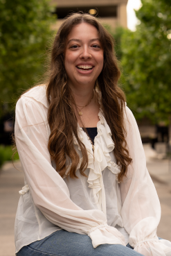 Smiling young woman with long brown hair sitting outdoors with greenery in the background, wearing a white blouse.