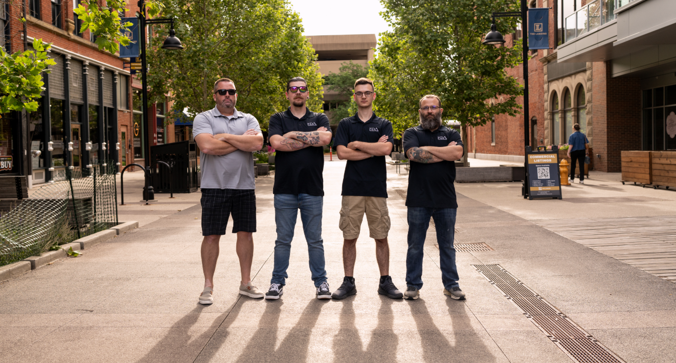 Four men standing with arms crossed on a city sidewalk, with buildings and trees in the background.