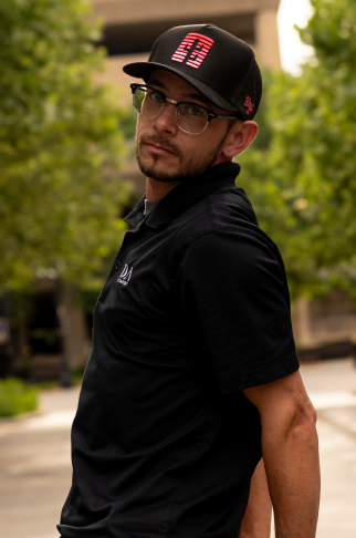 A young man wearing glasses, a black cap with pink and white writing, and a black polo shirt, standing outdoors with trees and a building in the background.