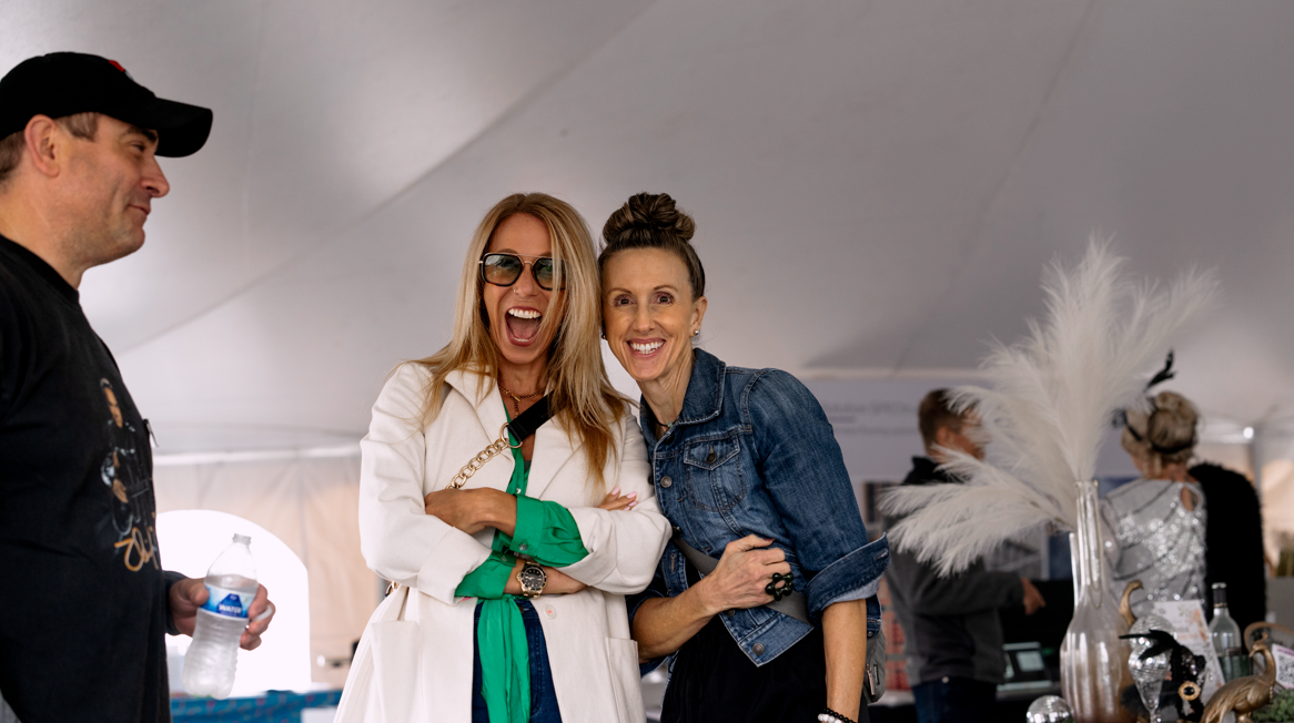 Three women smiling and talking at an indoor event, with two of them leaning close together. One woman is wearing sunglasses and a white coat, while the other is in a denim jacket. A man holding a water bottle is on their left. The background include