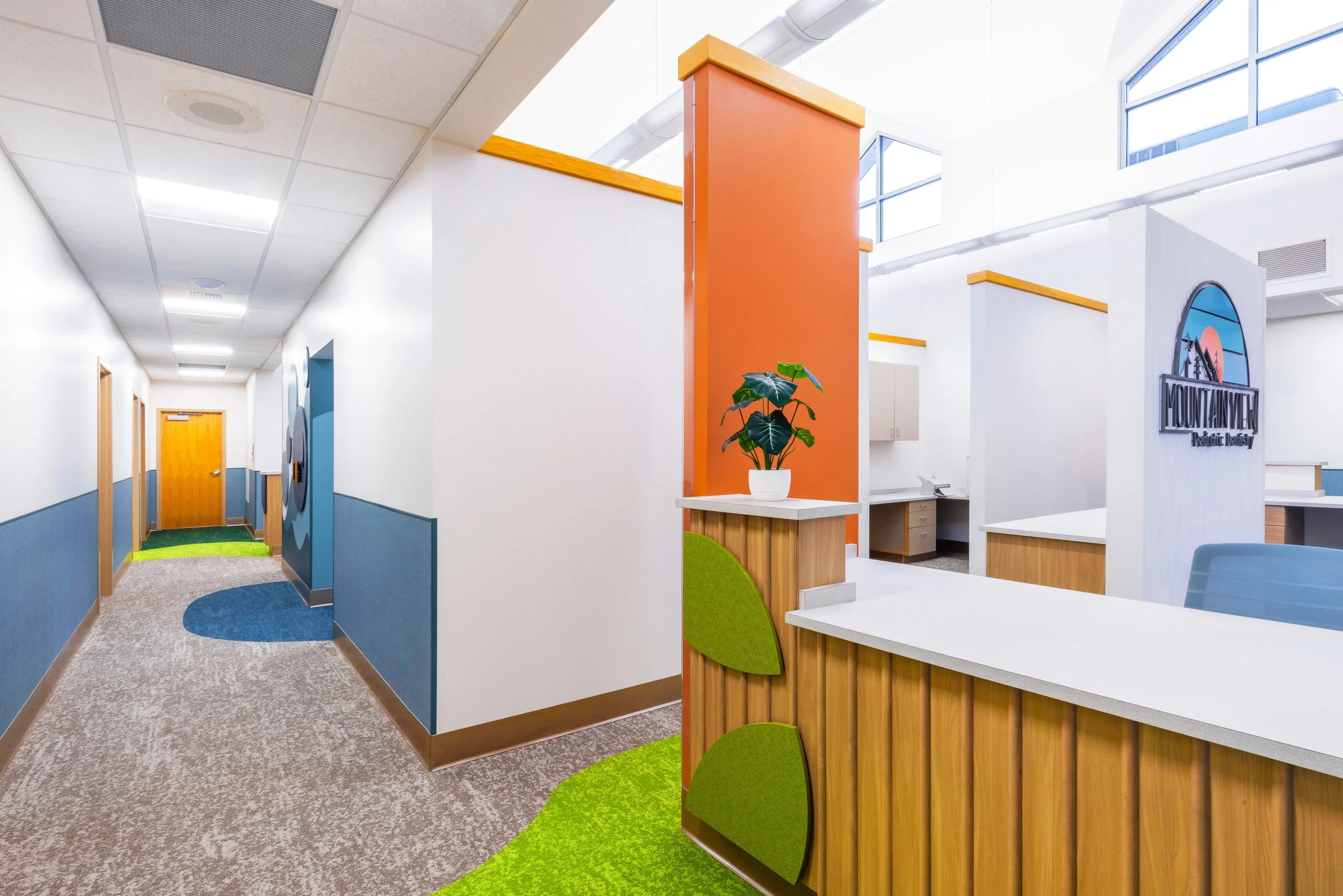 Interior view of a modern office or clinic with a reception desk, colorful walls, potted plant, and large windows.