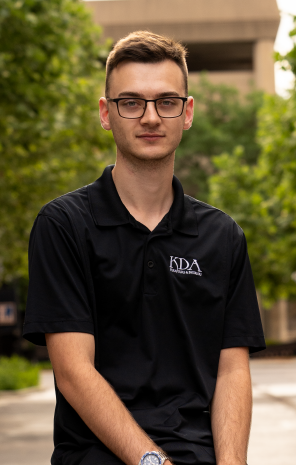 Young man wearing glasses and a black polo shirt with a logo, standing outdoors in front of greenery and a building.