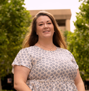A smiling woman with long hair standing outdoors in front of a building and trees.