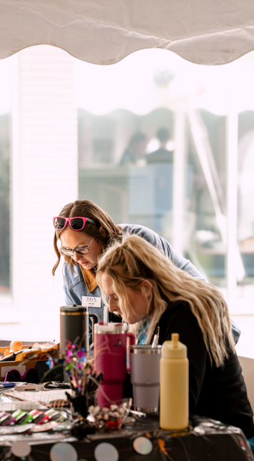 Woman and girl sitting at a table with makeup and beauty products, outdoors under a canopy