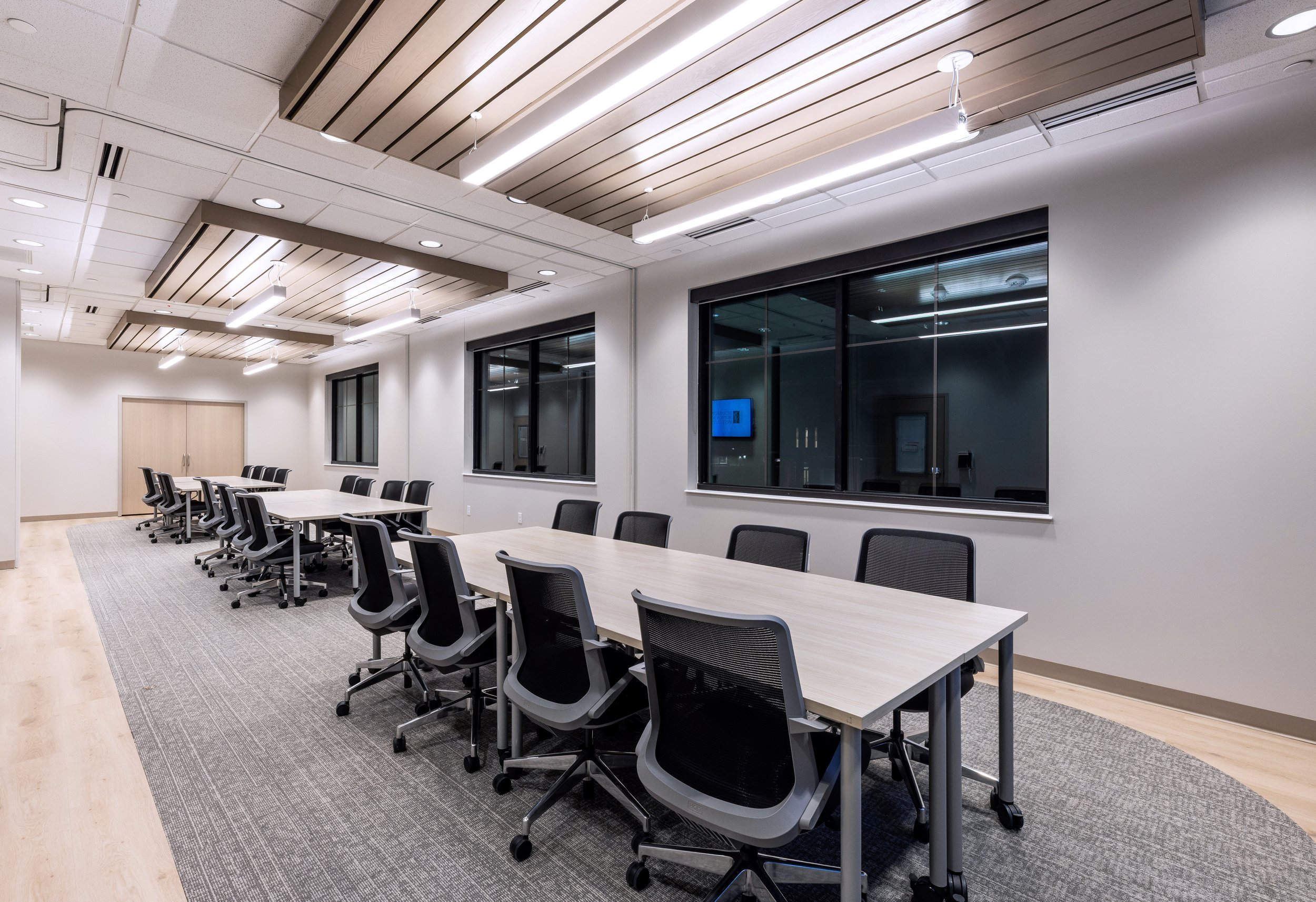 Empty conference room with long tables and black office chairs, large windows with blinds, and modern ceiling lights in a professional office setting.
