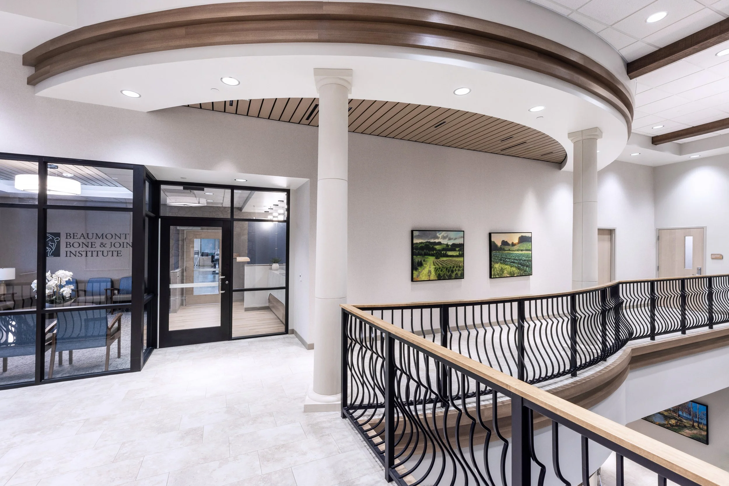 Interior view of an office or clinic lobby with white walls, a wooden ceiling with lighting, framed landscape paintings on the wall, a black metal railing along a staircase, and an entrance to Beaumont Bone & Joint Institute.
