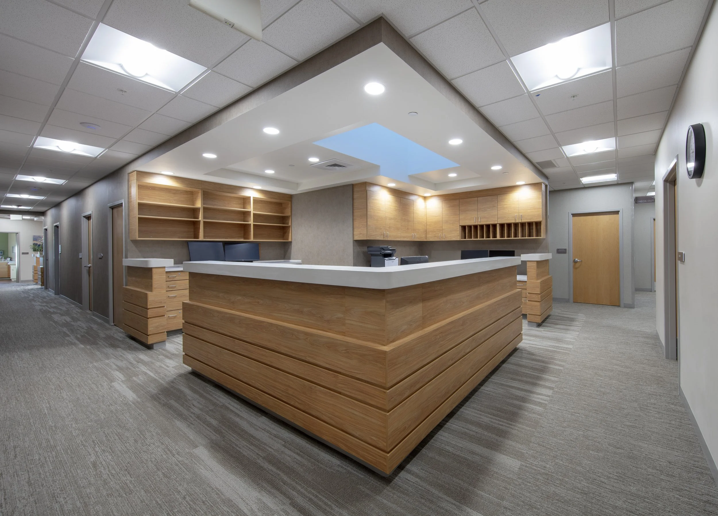 Reception area with woodgrain furniture, open shelving, a curved front desk, and overhead lighting in a modern office.