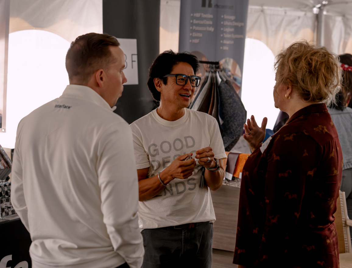 Three people engaged in a conversation inside a tent or event space. The person in the middle is wearing glasses and a white T-shirt that says 'GOOD DEEDS' and is smiling, while the person on the right, with curly hair and a floral-patterned jacket, is gesturing with her hand. The person on the left has short hair and is wearing a white shirt.
