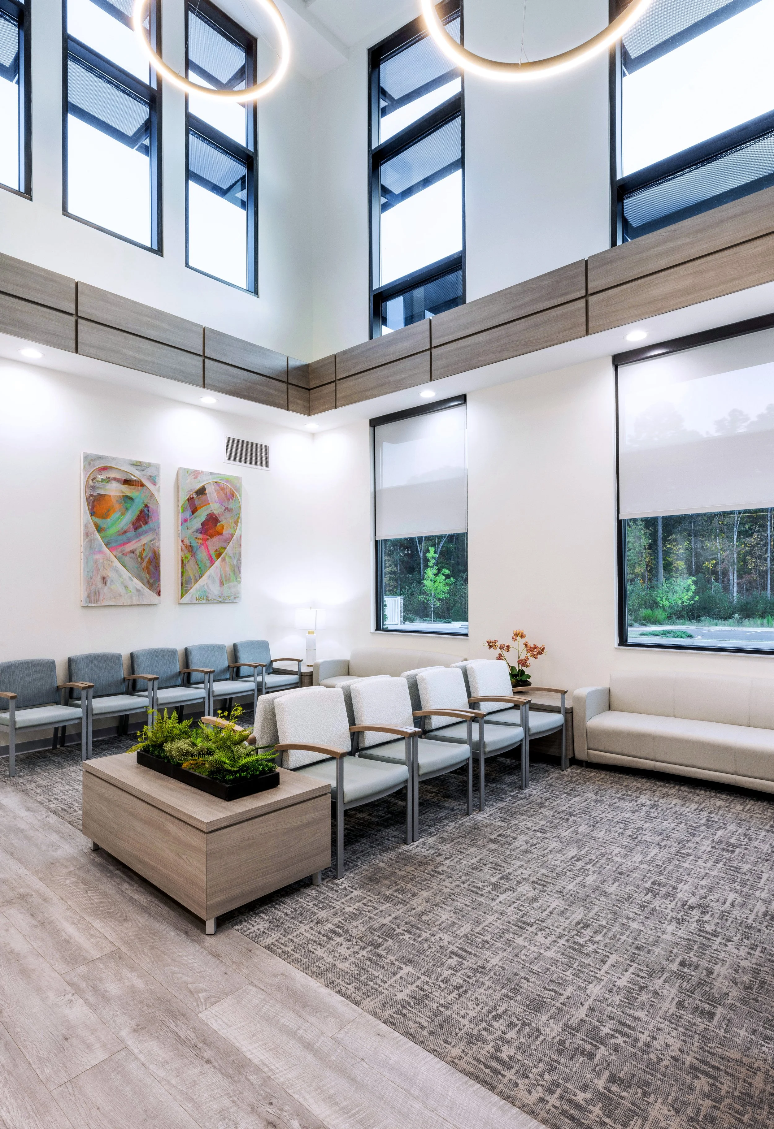 A modern waiting room with rows of chairs, a large window, abstract heart artwork on the wall, a rug, and a rectangular planter with green plants on a table. The room has high ceilings with large circular lights, white walls, and a wood-paneled mezzanine.