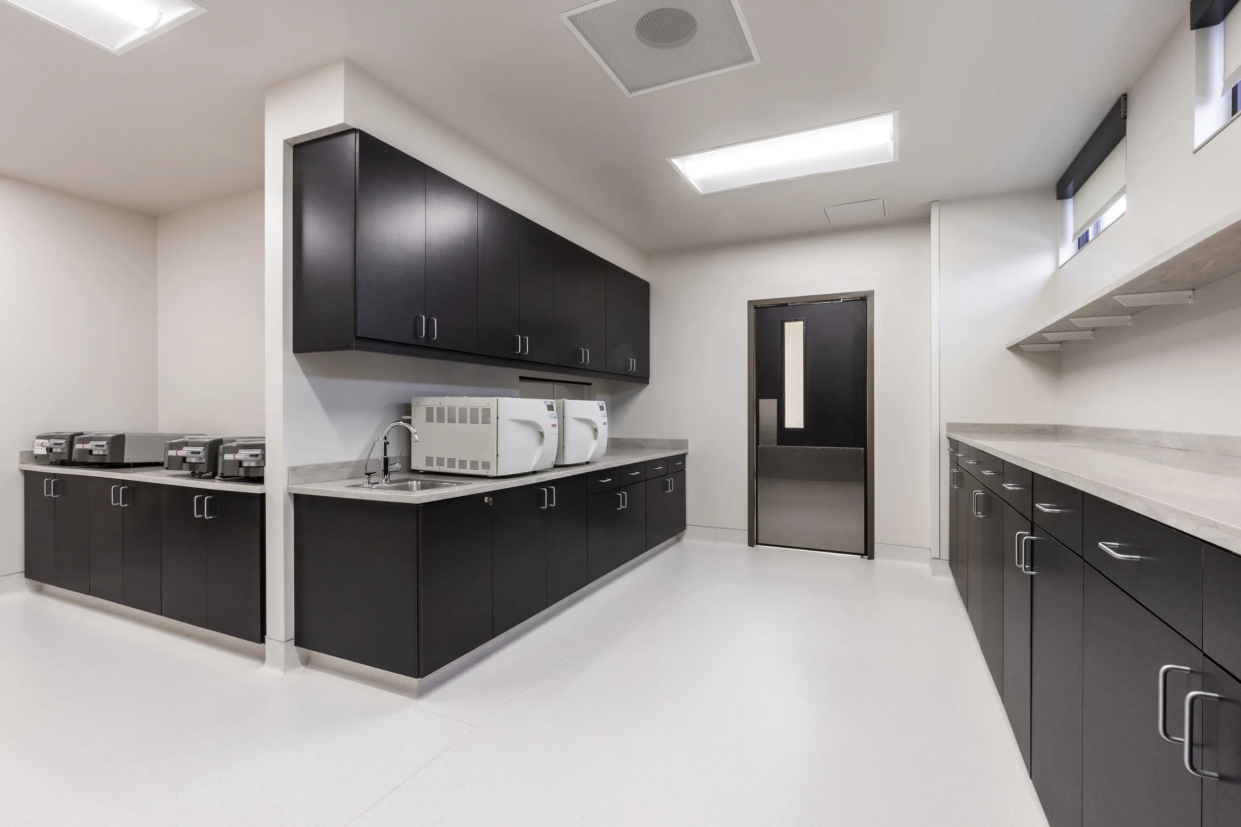 A modern kitchen with black cabinets, a light countertop, and a white wall. There are small appliances on the counter, including microwaves and toasters. The ceiling has fluorescent lights and there is a black door at the back.