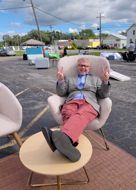 A smiling man with glasses and gray hair making peace signs with both hands while sitting in a light pink armchair outdoors. There is a small table in front of him with his feet resting on it. The background includes a parking lot, utility poles, some houses, and a partly cloudy sky.