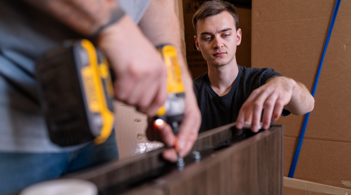 Two young men assembling furniture, one is using a cordless drill to secure screws, the other is holding a wooden piece steady, surrounded by cardboard boxes.