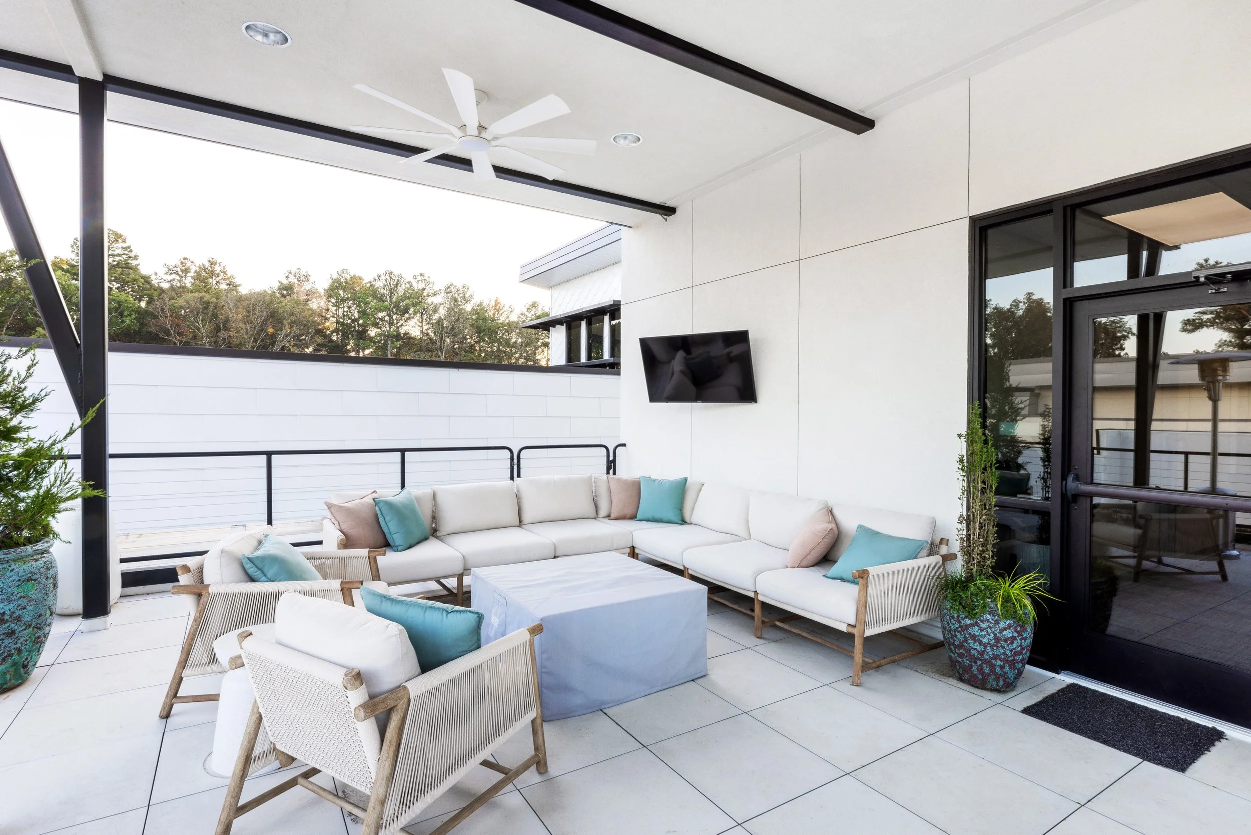 An outdoor patio area with white tiled flooring, a white sectional sofa with pastel pillows, and a small coffee table. There are large potted plants, black railings, a wall-mounted TV, and a ceiling fan across a scenic view of trees.