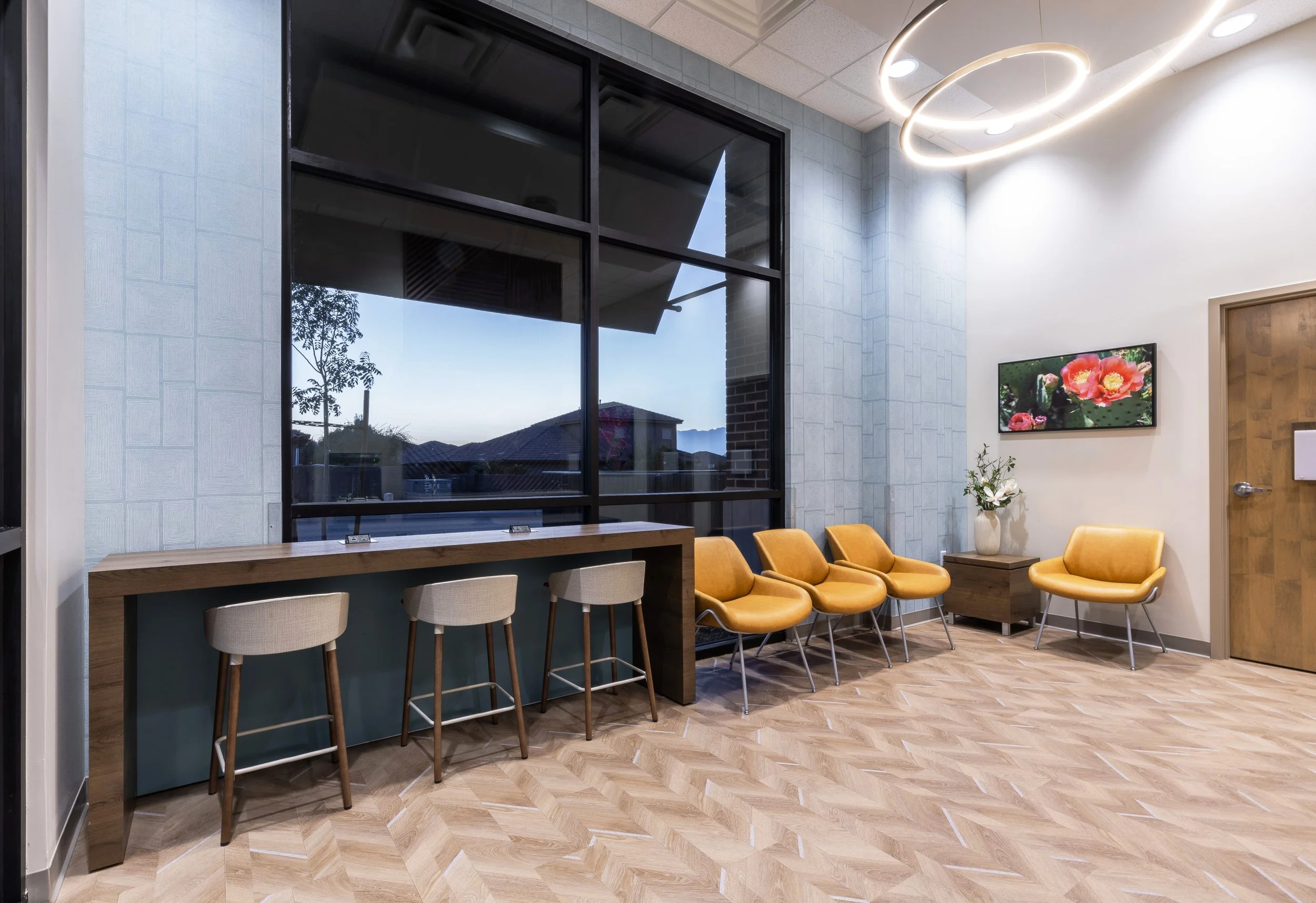 Waiting room or reception area with a row of orange chairs, a small table with flowers, a wall-mounted TV displaying flowers, and large windows showing an outdoor scene with houses and trees.
