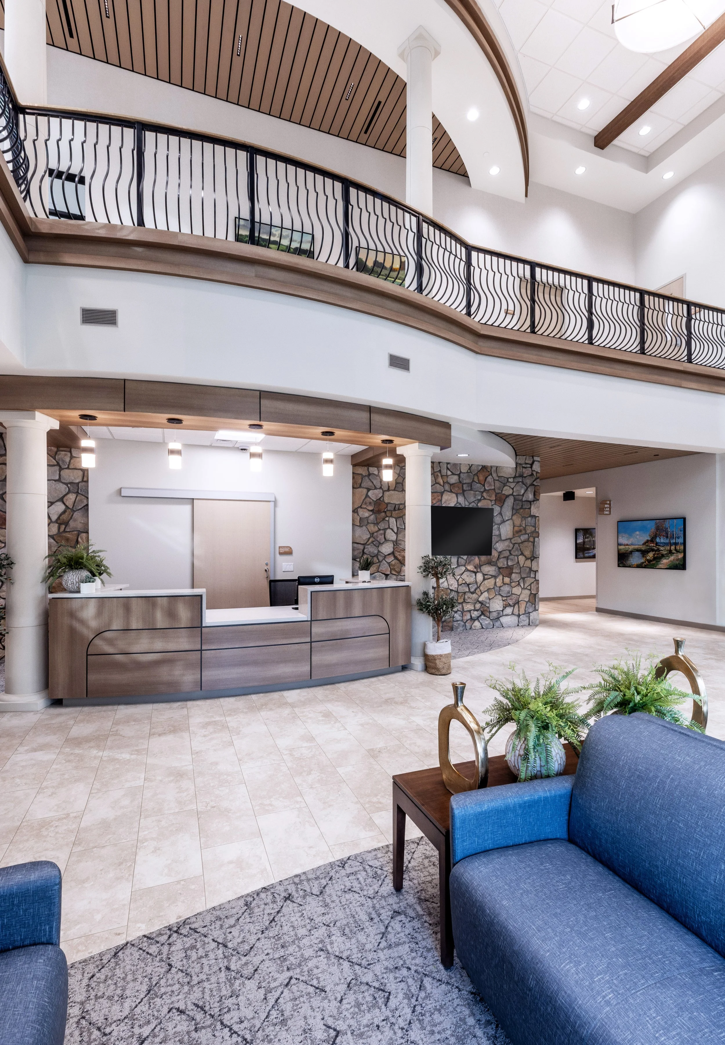 Lobby area with a reception desk, stone accent wall, and comfortable seating. The space features a double-height ceiling with recessed lighting, wood ceiling accents, and a balcony with a wavy black metal railing.