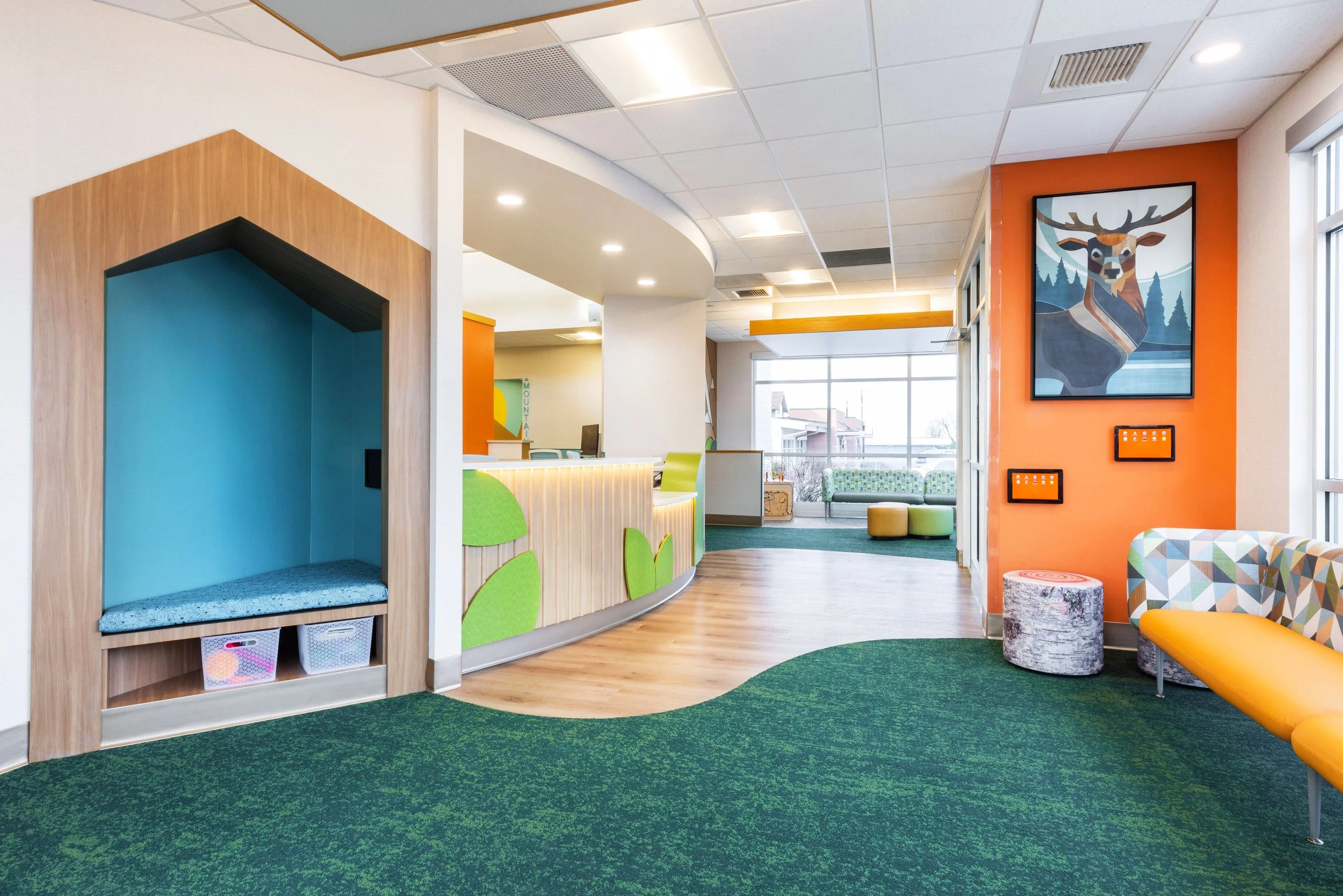 Interior of a colorful children's waiting room with green and orange accents, seating, and large windows.