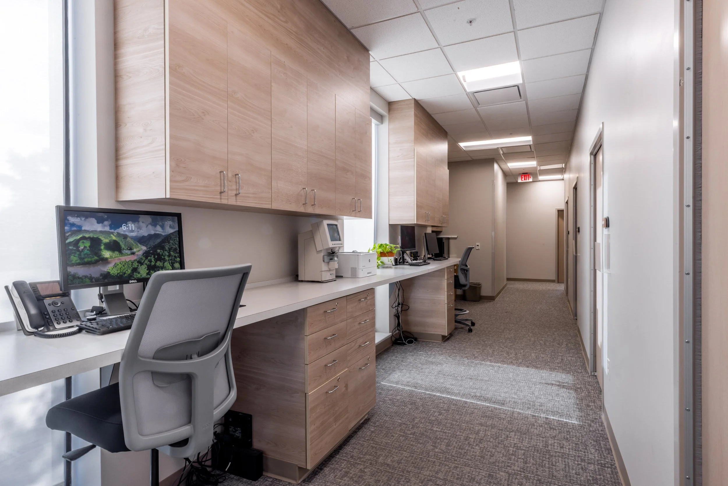 An office space with desks, computers, cabinets, and office chairs, with a corridor and doors in the background.