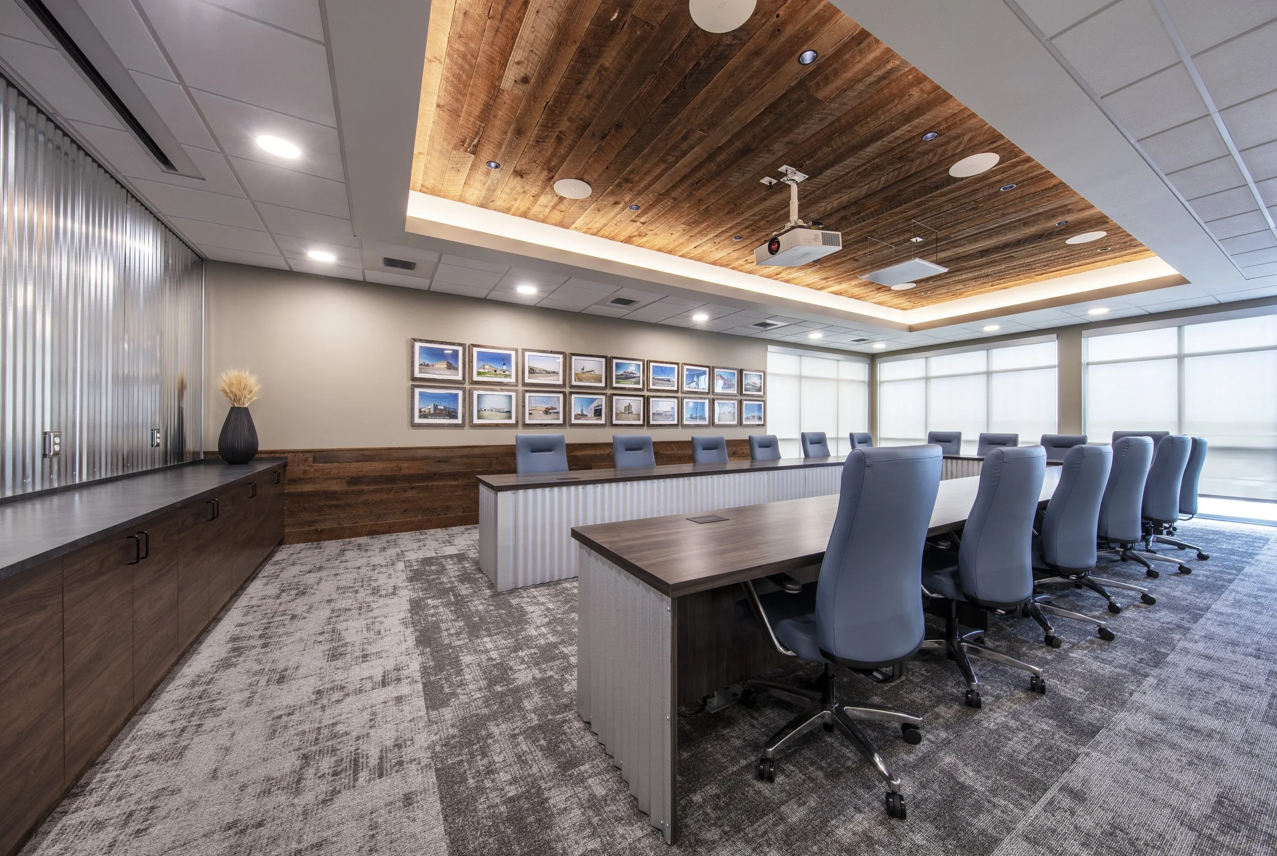 A modern conference room with a long table, blue office chairs, framed photos on the wall, a ceiling with wood paneling, recessed lighting, and large windows with blinds.