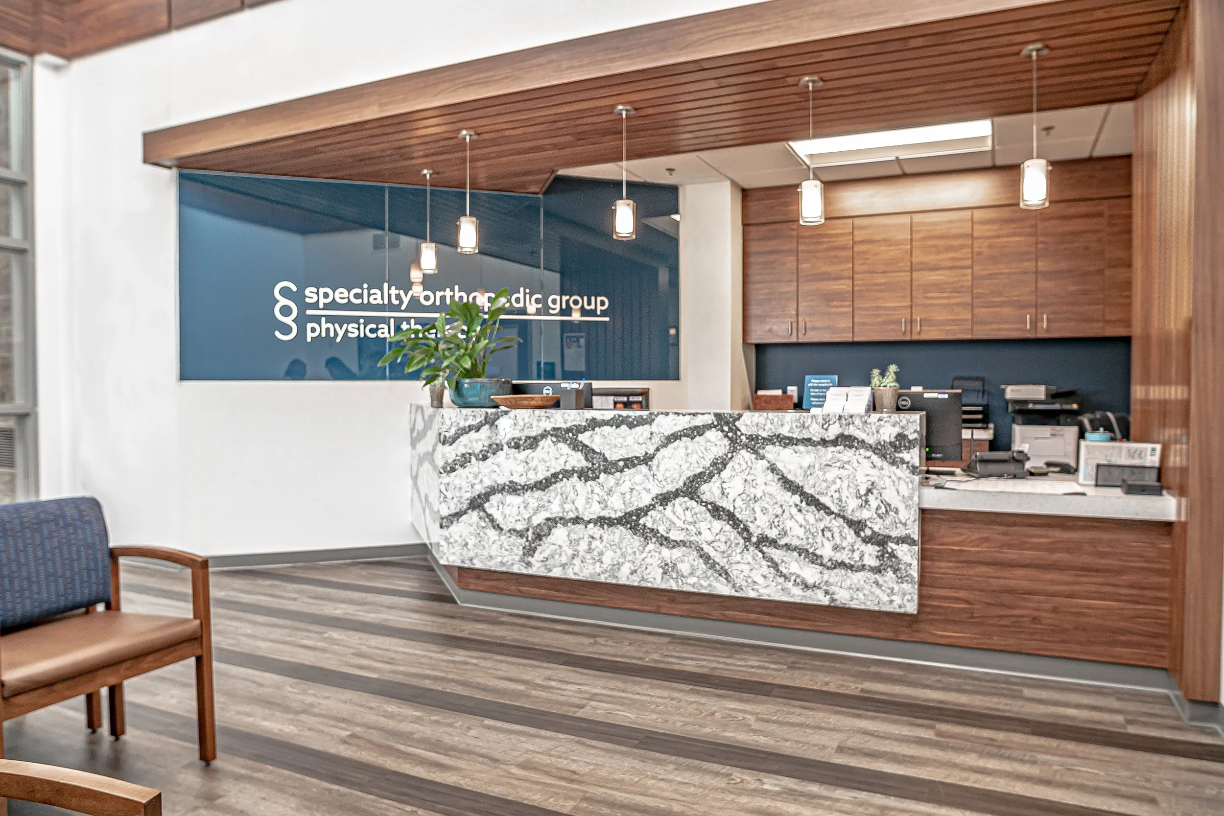Reception desk with a marble countertop, a sign on the wall for a specialty orthopedic group, wooden cabinets, hanging pendant lights, chairs, and office supplies in an office lobby.