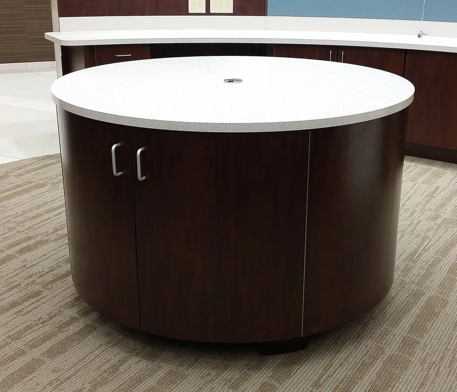 A circular kitchen island with a white countertop and dark wooden cabinet base located in an office or commercial space.
