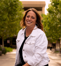 A smiling woman with shoulder-length hair standing outdoors with trees and a building in the background.