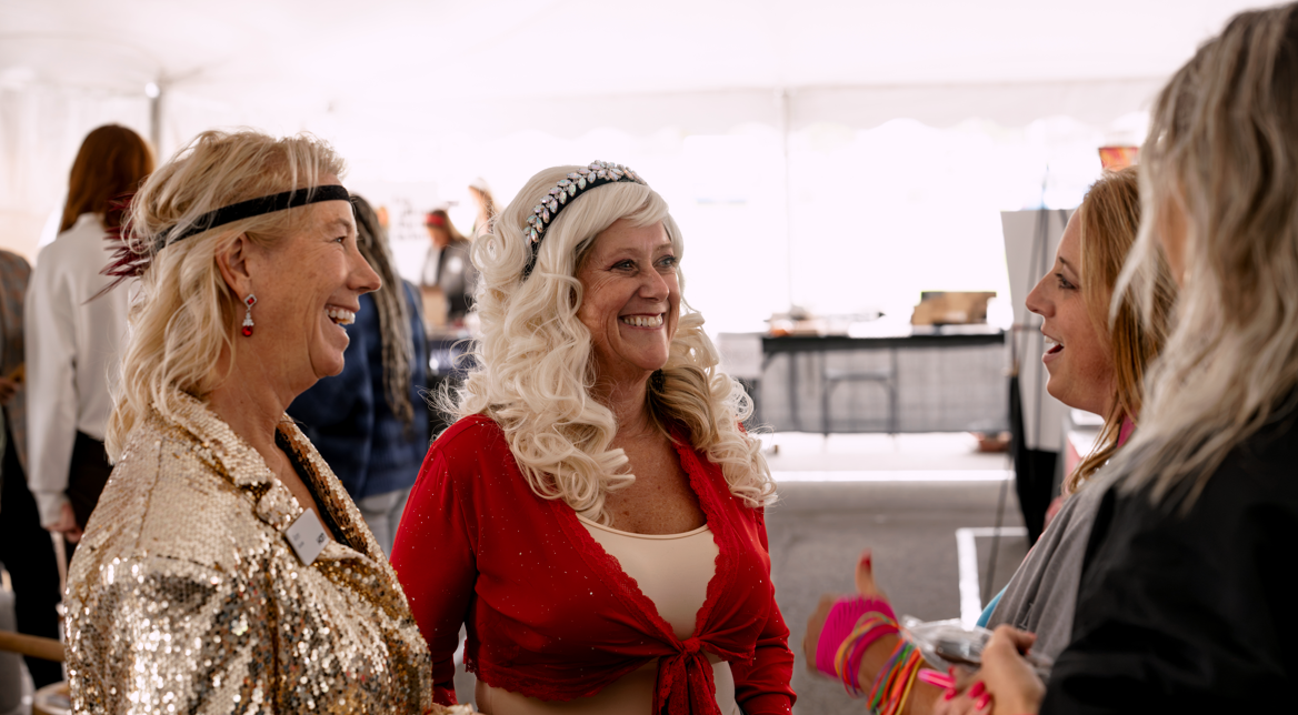 Four women chatting and smiling at an indoor event, with a table and other people in the background.