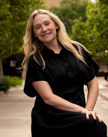 A young woman with long blonde hair smiling outdoors in a black dress, standing on a city street with green trees in the background.
