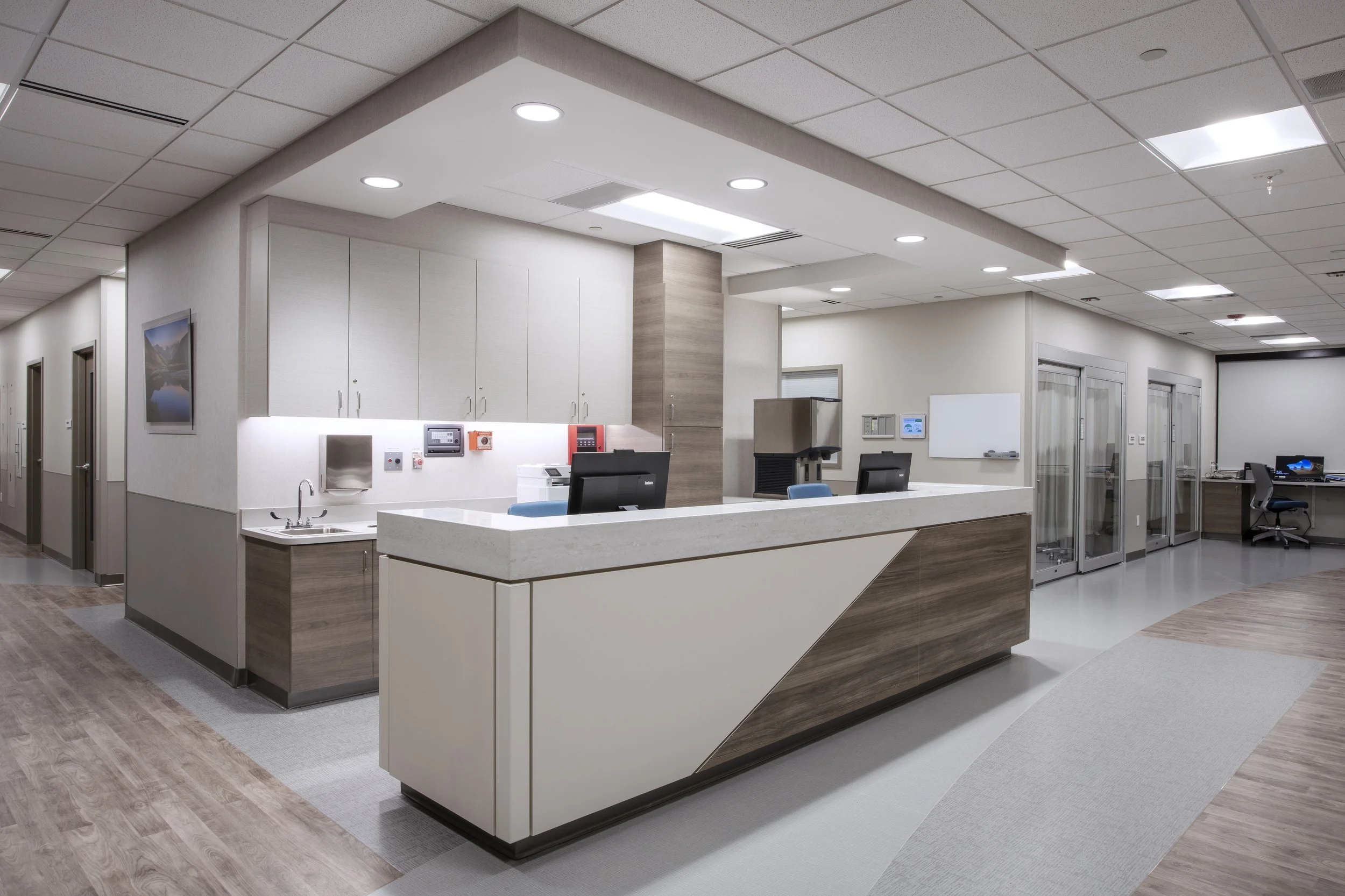 Modern hospital reception area with white and light wood cabinetry, computer stations, whiteboard, and glass elevators, illuminated by ceiling lights.