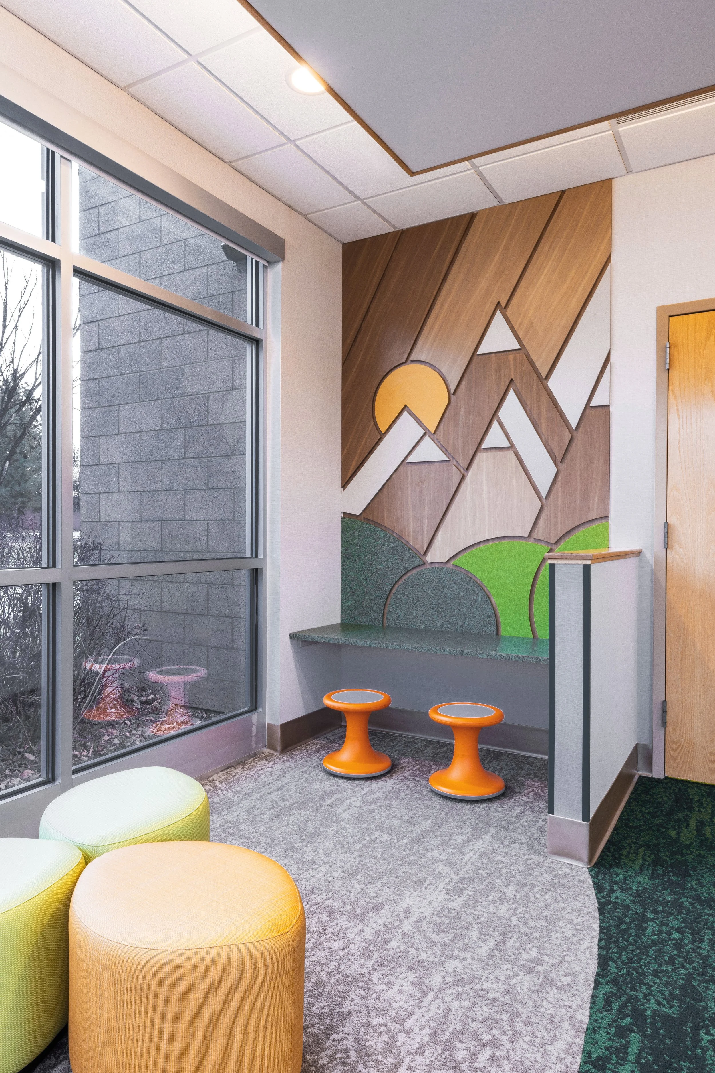 Interior of a room with large window, colorful ottomans, a wall mural of mountains with a sunset, and a small counter with orange stools.