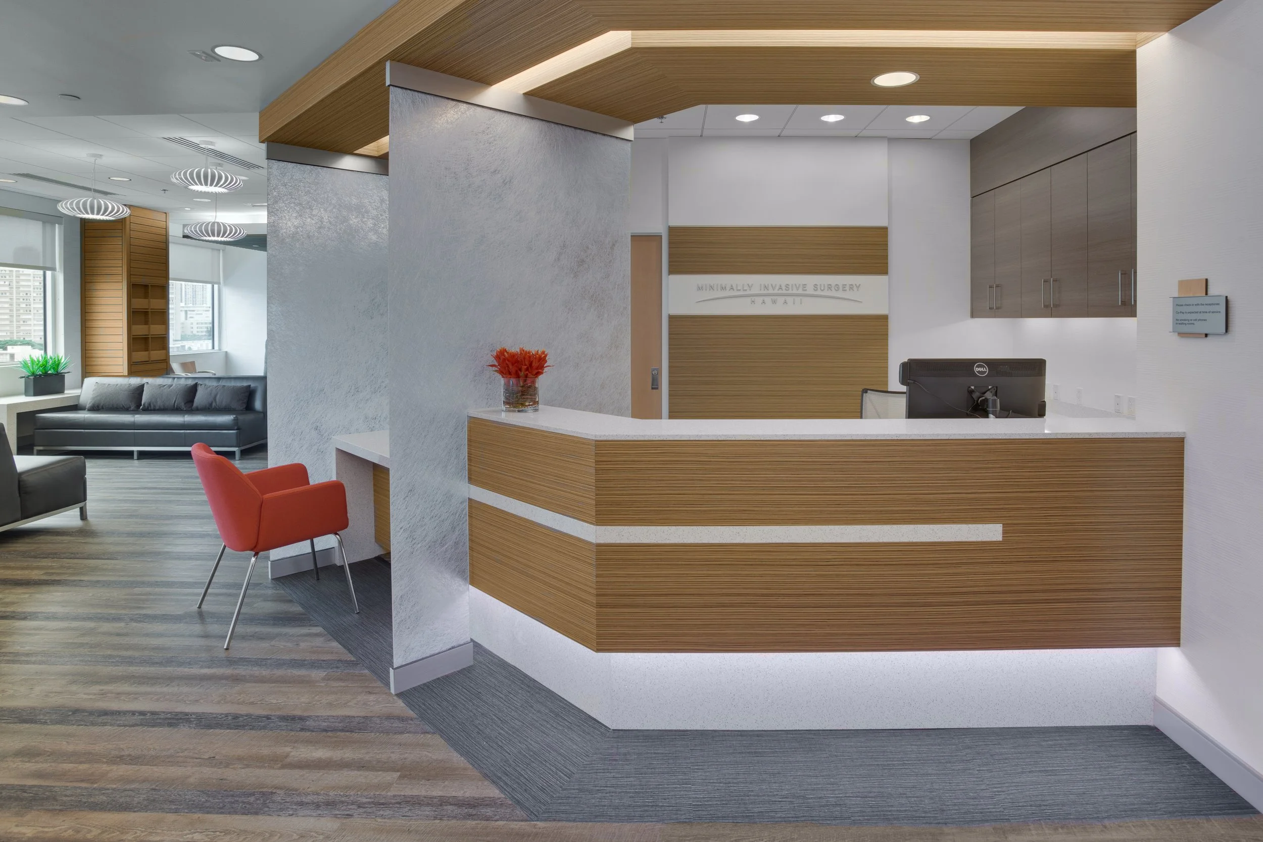 Modern medical office waiting area with wooden accents, a reception desk, a black leather sofa, a red chair, and decorative lighting in a high-rise building.