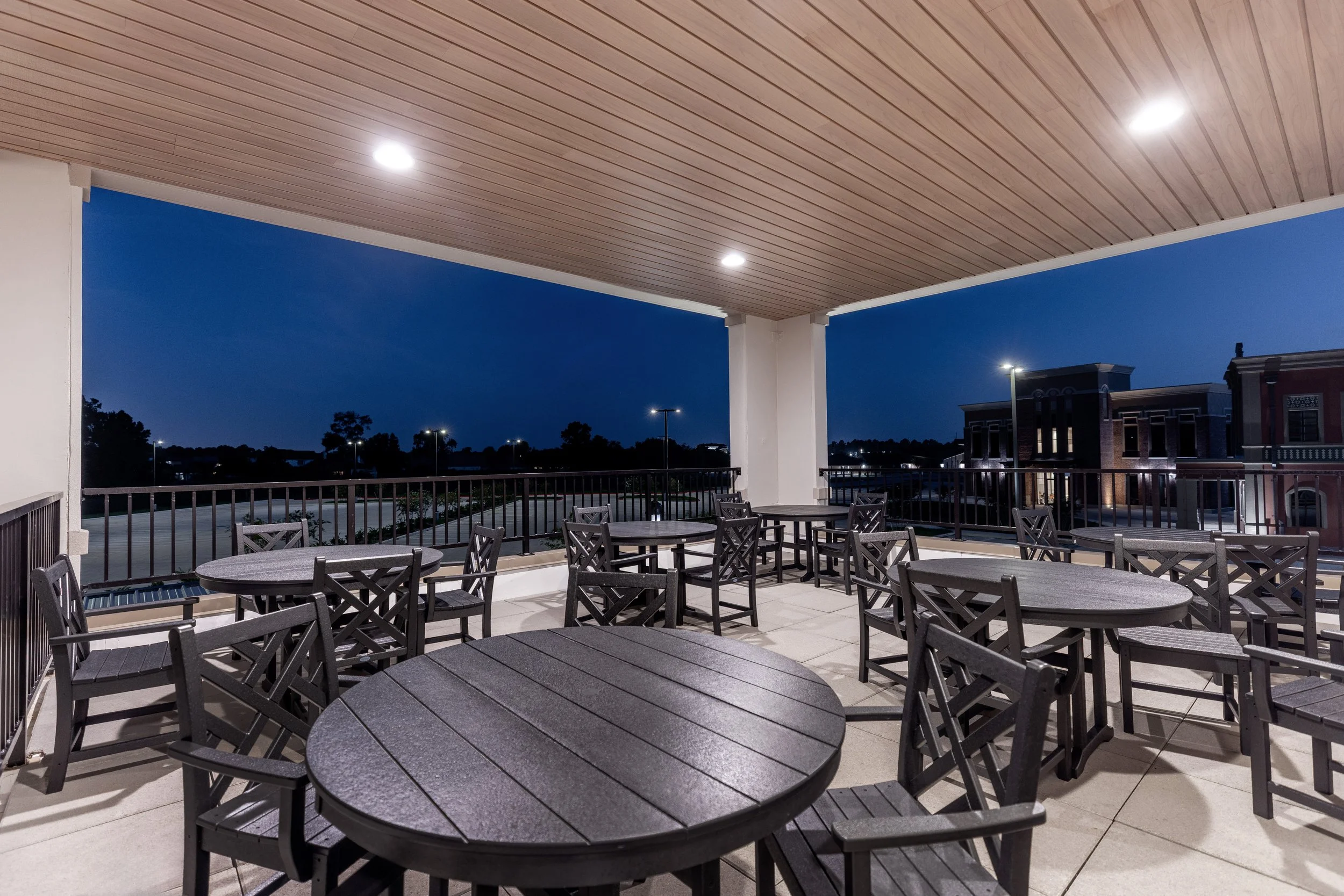 Night view of an outdoor patio with round tables and chairs, overhead ceiling with recessed lighting, railing, and a cityscape with buildings and trees in the background.