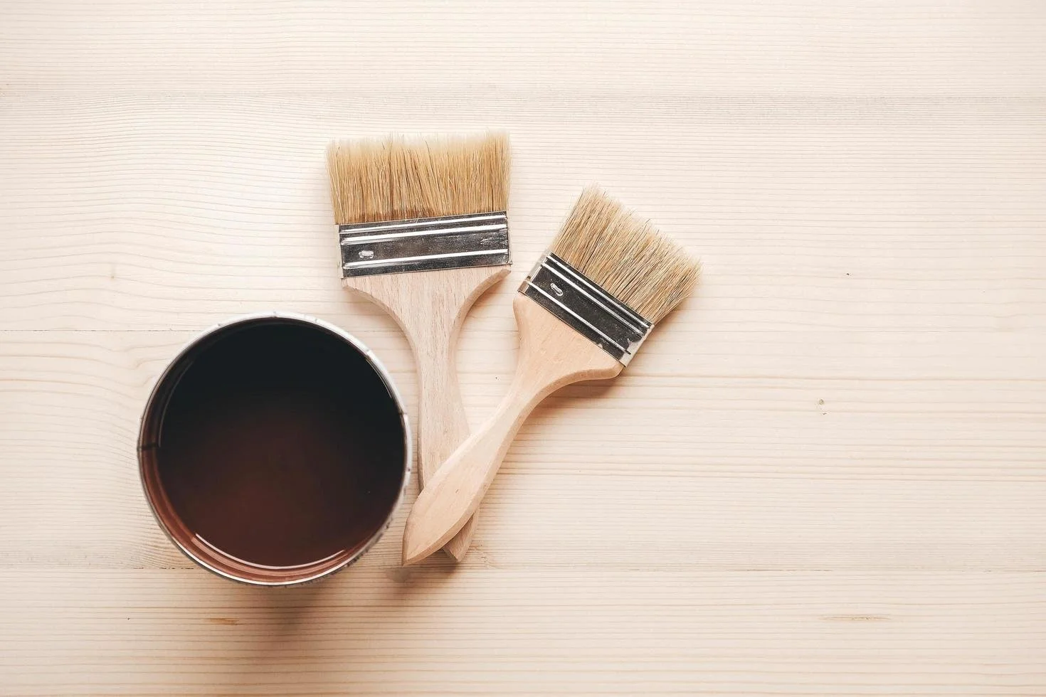 Two paintbrushes and a can of dark paint on a light wooden surface.