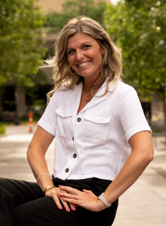 A woman with blonde hair smiling outdoors in a white button-up shirt and black pants, sitting on a park bench with trees in the background.