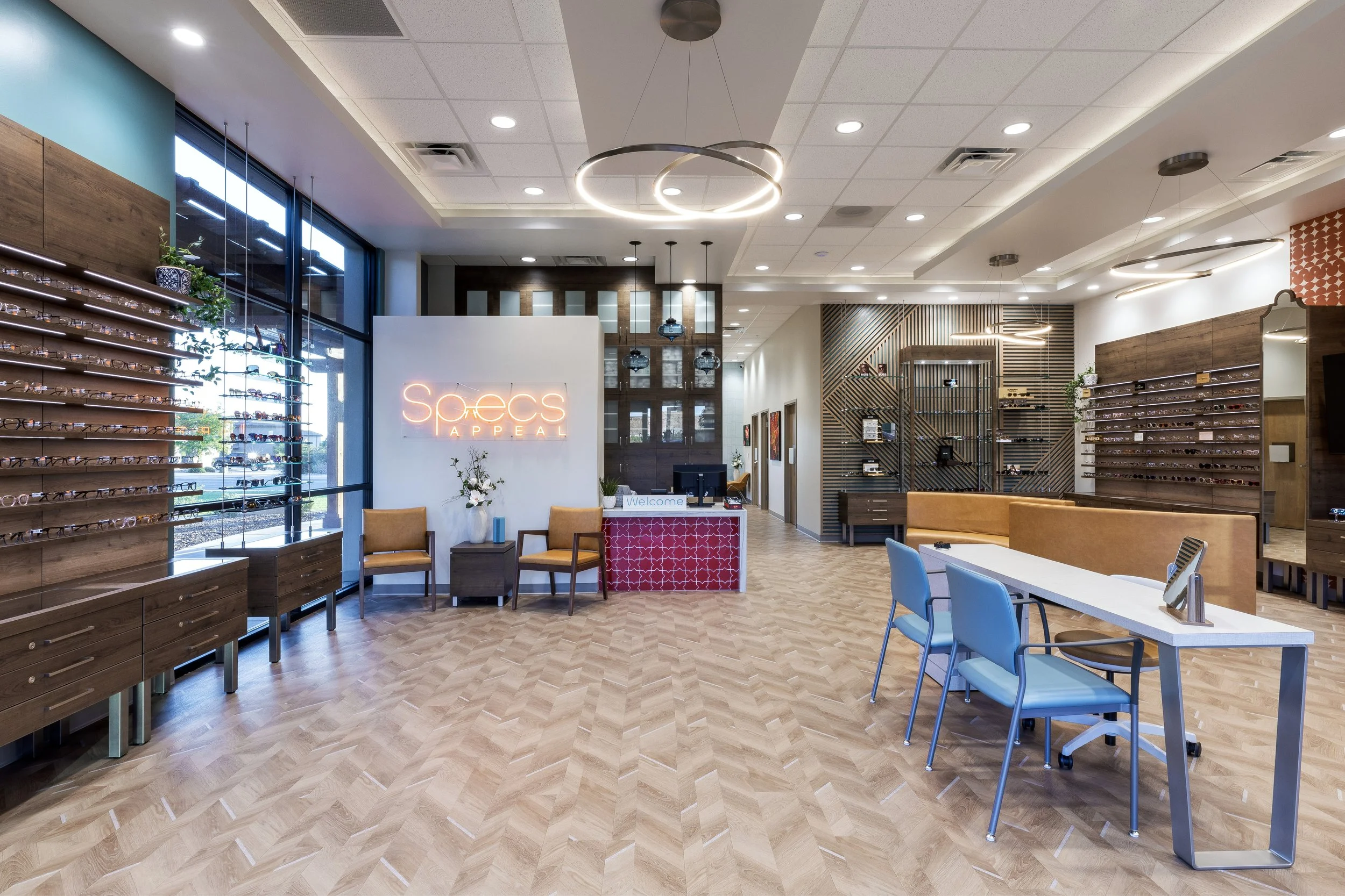 Interior of a modern eyewear retail store with eyeglasses on wooden display shelves, a seating area with chairs and tables, large windows, and a neon sign that reads 'Specs Appeal' behind the reception desk.