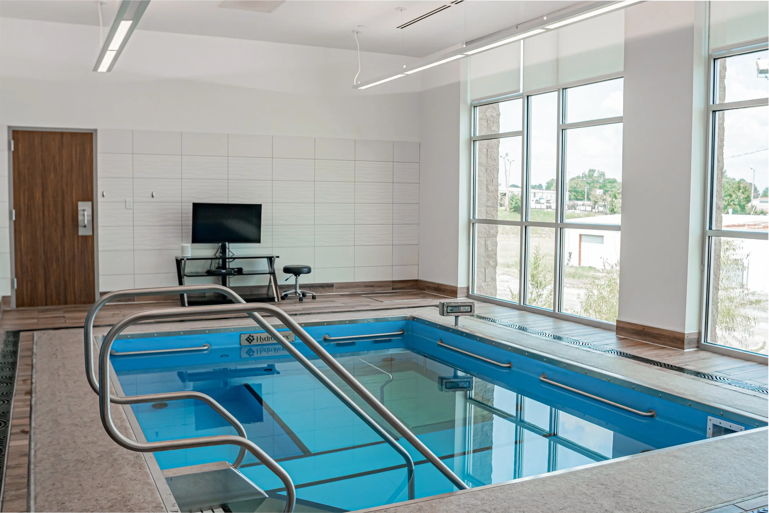 Indoor swimming pool with a whirlpool and large windows providing natural light and a view of the outside area.
