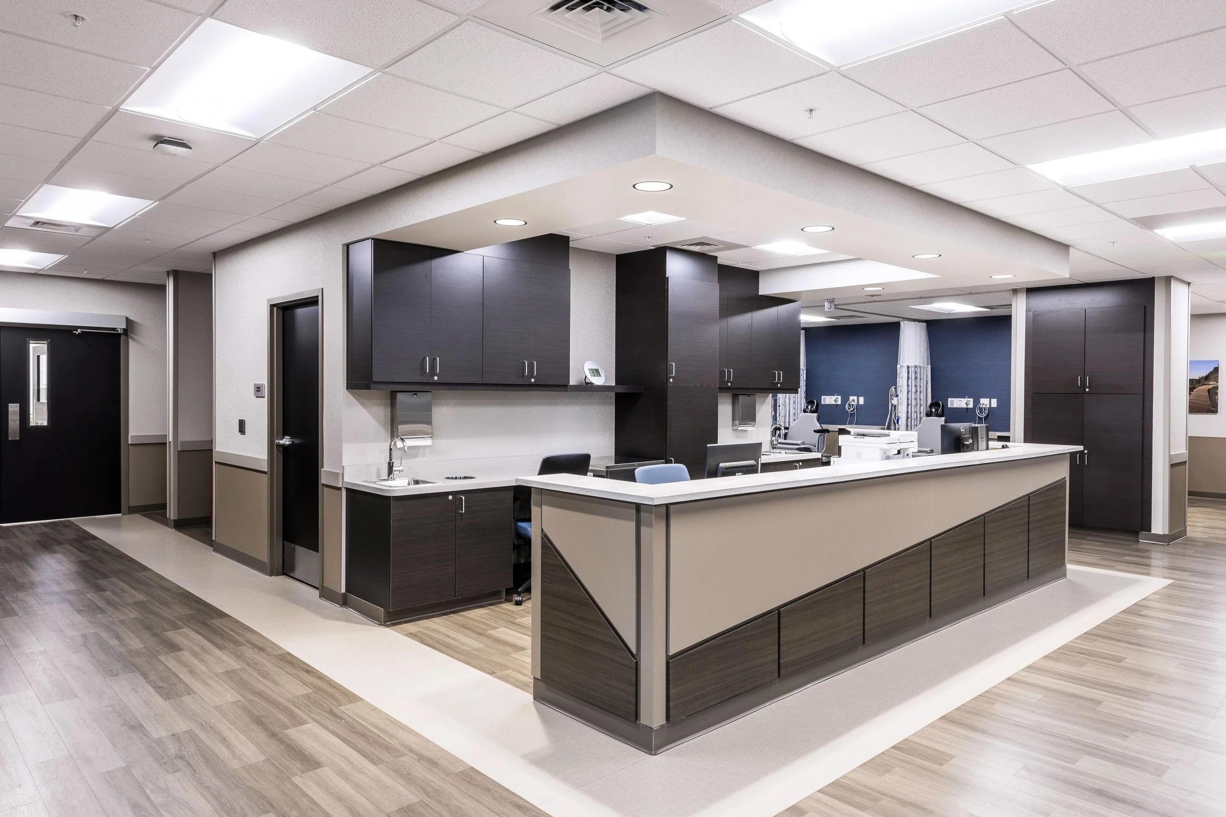 A hospital nurse station with dark wood cabinets, a white countertop, a computer, and medical equipment. The room has a drop ceiling with fluorescent lights and wood-like flooring.