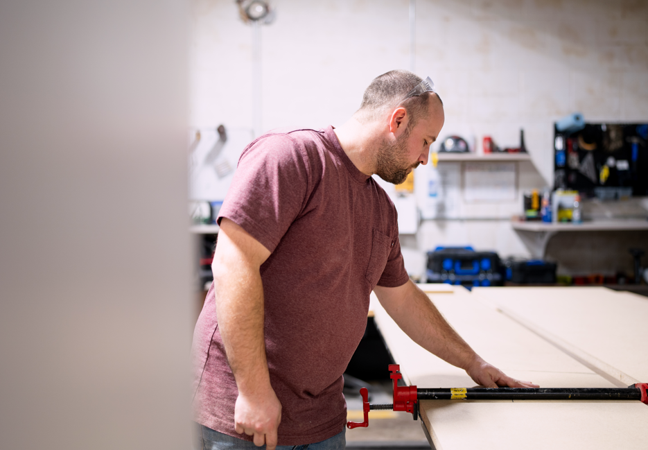 A man in a maroon t-shirt working on a woodworking project in a workshop. He is using a clamp to secure a piece of wood on a workbench.