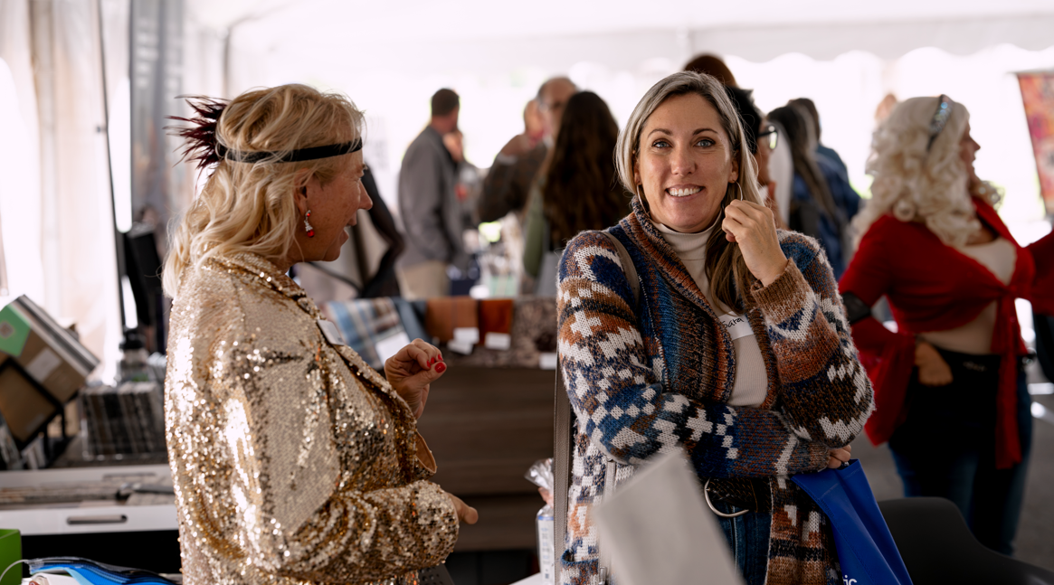 A woman with long hair wearing a patterned cardigan is smiling with her hand near her face, standing at an indoor event. Next to her, another woman with blonde hair, dressed in a gold sequined jacket, is talking. In the background, multiple people ar