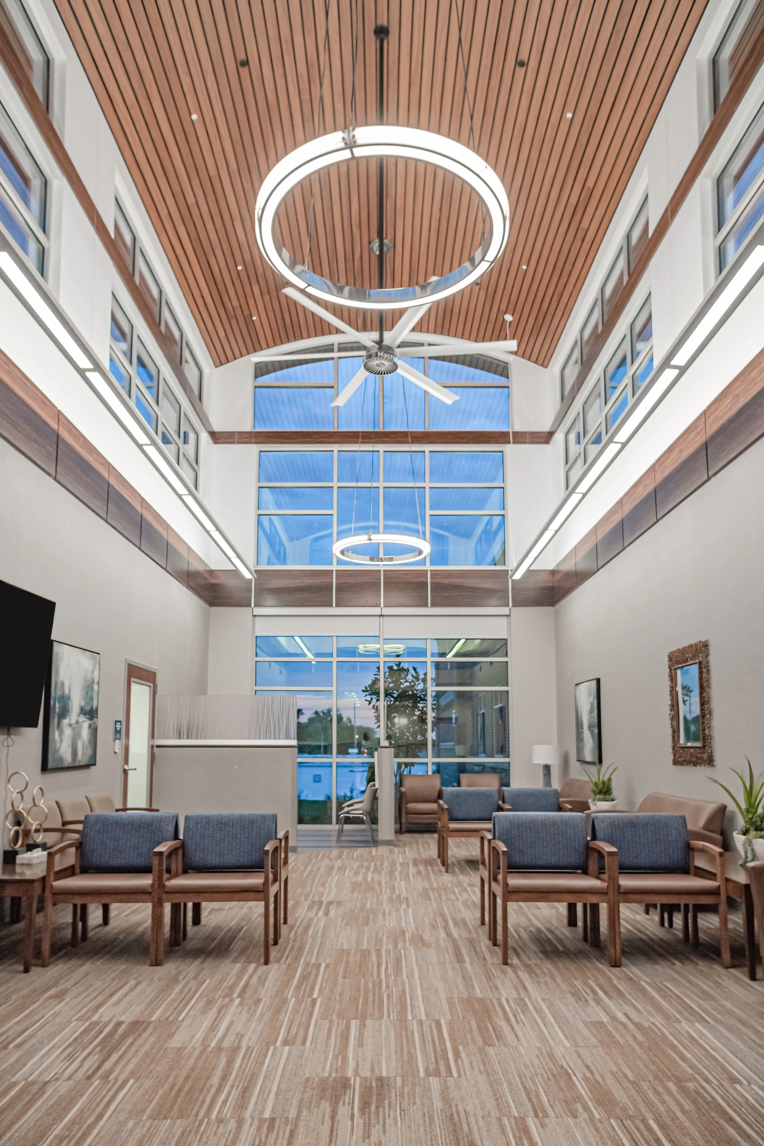 A spacious, modern lobby with wooden ceiling, large windows, circular light fixtures, ceiling fan, and seating area with chairs, sofas, and artwork on the walls.